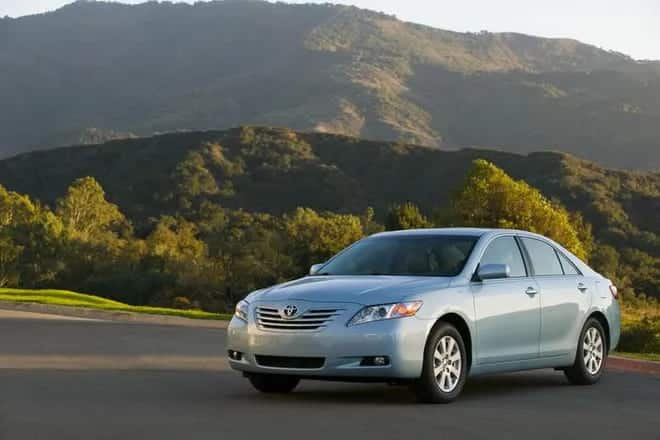 A view of a silver Toyota camry driving on a road with mountains and trees in the background